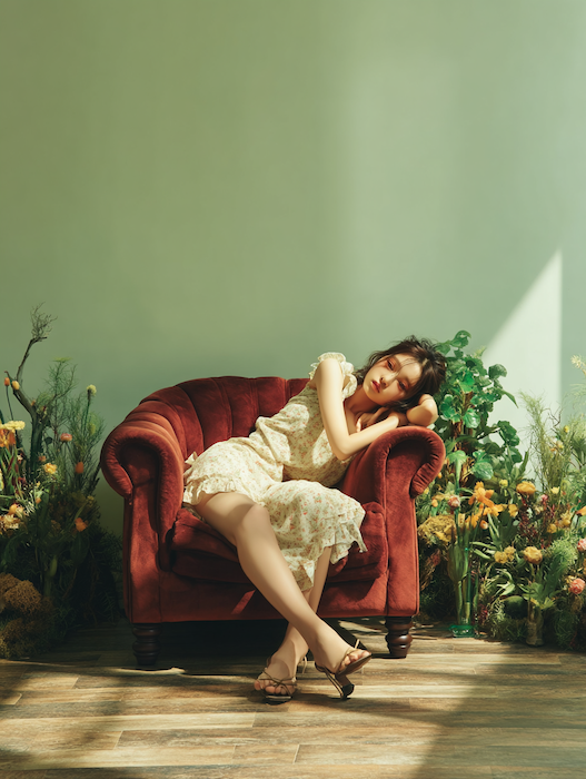 The virtual artist in a vintage floral dress draped across a crimson velvet armchair against a sage green wall, surrounded by plants, in warm afternoon light